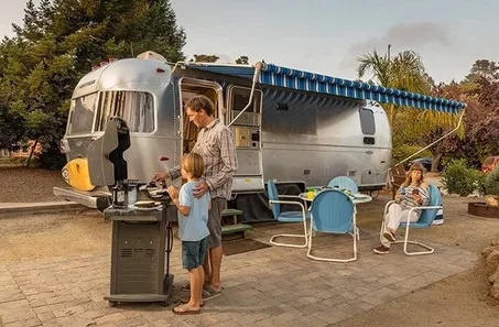A family cooking outdoors near an RV with a side arm awning.
